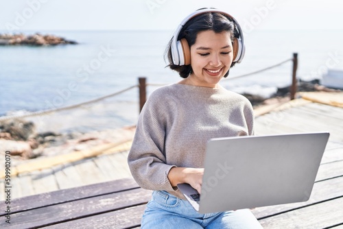 Young woman using laptop and headphones sitting on bench at seaside © Krakenimages.com