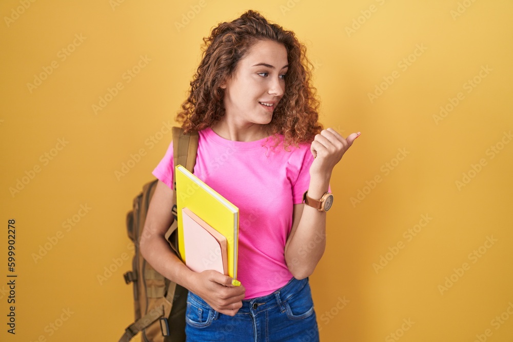 Young caucasian woman wearing student backpack and holding books smiling with happy face looking and pointing to the side with thumb up. Young caucasian woman wearing student backpack and holding books smiling with happy face looking and pointing to the side with thumb up.
