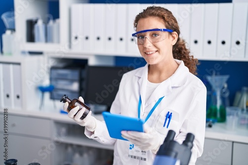 Young beautiful hispanic woman scientist using touchpad holding bottle at laboratory