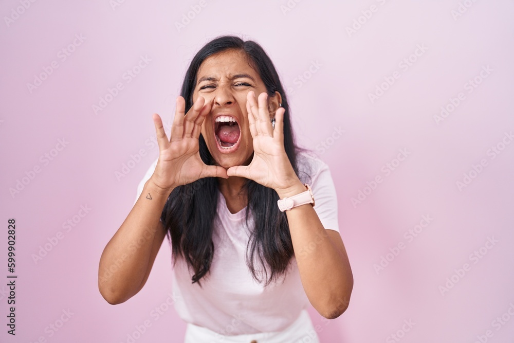 Young hispanic woman standing over pink background shouting angry out loud with hands over mouth Young hispanic woman standing over pink background shouting angry out loud with hands over mouth