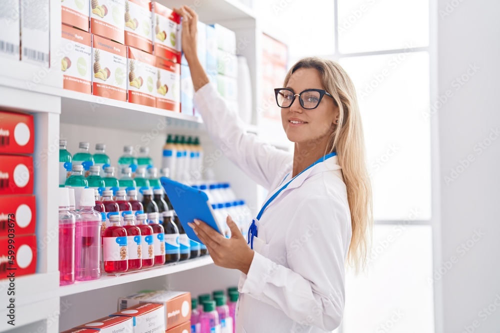 Young blonde woman pharmacist using touchpad holding product on shelving at pharmacy