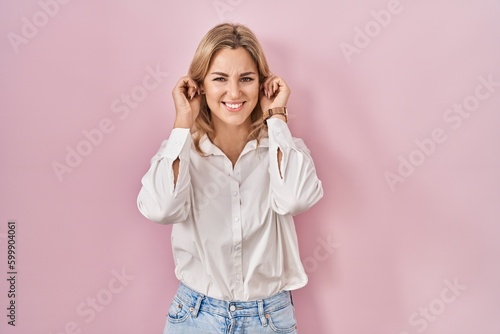 Young caucasian woman wearing casual white shirt over pink background covering ears with fingers with annoyed expression for the noise of loud music. deaf concept.