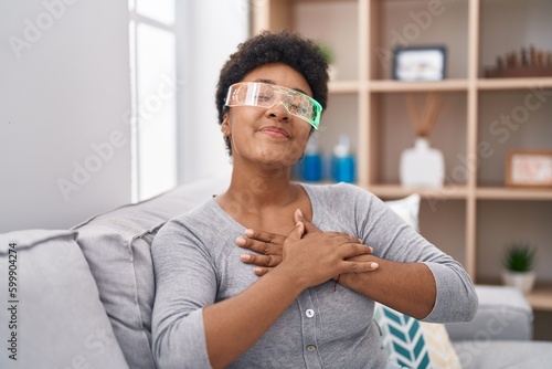 Young african american woman wearing virtual reality glasses sitting on the sofa smiling with hands on chest, eyes closed with grateful gesture on face. health concept.