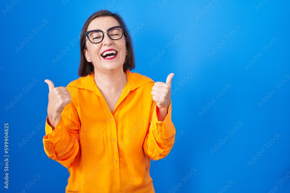 Middle age hispanic woman wearing glasses standing over blue background success sign doing positive gesture with hand, thumbs up smiling and happy. cheerful expression and winner gesture. Middle age hispanic woman wearing glasses standing over blue background success sign doing positive gesture with hand, thumbs up smiling and happy. cheerful expression and winner gesture.
