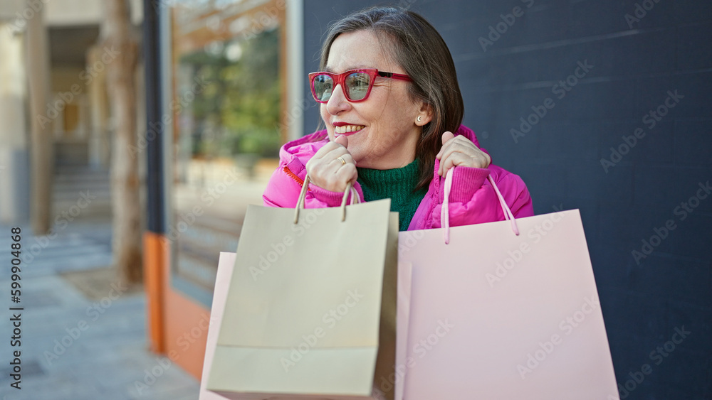 Mature hispanic woman with grey hair smiling going shopping holding bags at street