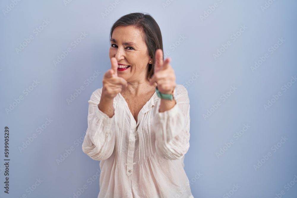 Middle age hispanic woman standing over blue background pointing fingers to camera with happy and funny face. good energy and vibes. Middle age hispanic woman standing over blue background pointing fingers to camera with happy and funny face. good energy and vibes.