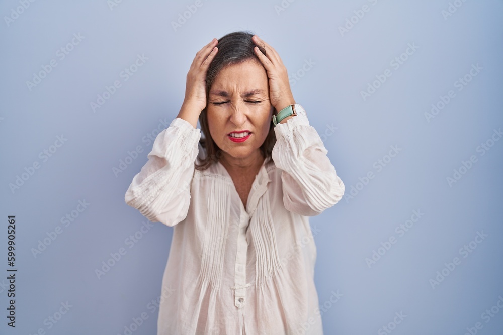 Middle age hispanic woman standing over blue background suffering from headache desperate and stressed because pain and migraine. hands on head. Middle age hispanic woman standing over blue background suffering from headache desperate and stressed because pain and migraine. hands on head.