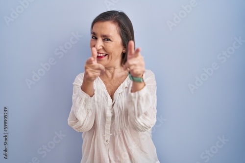 Middle age hispanic woman standing over blue background pointing fingers to camera with happy and funny face. good energy and vibes.