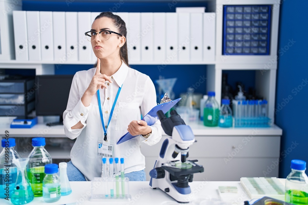 Young brunette woman working at scientist laboratory thinking concentrated about doubt with finger on chin and looking up wondering