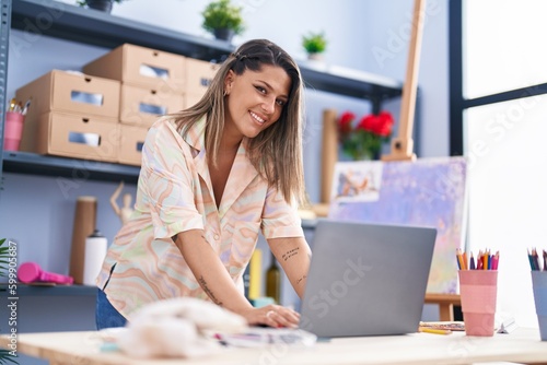 Young hispanic woman artist smiling confident using laptop at art studio