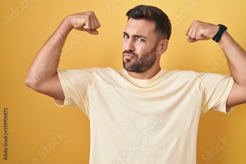 Handsome hispanic man standing over yellow background showing arms muscles smiling proud. fitness concept.