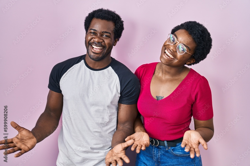 Obraz premium Young african american couple standing over pink background smiling cheerful with open arms as friendly welcome, positive and confident greetings