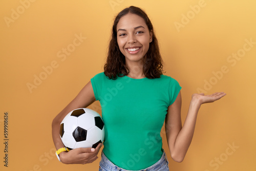 Young hispanic woman holding ball smiling cheerful presenting and pointing with palm of hand looking at the camera. © Krakenimages.com