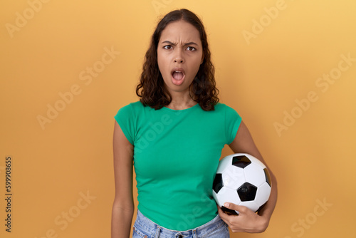 Young hispanic woman holding ball in shock face, looking skeptical and sarcastic, surprised with open mouth © Krakenimages.com