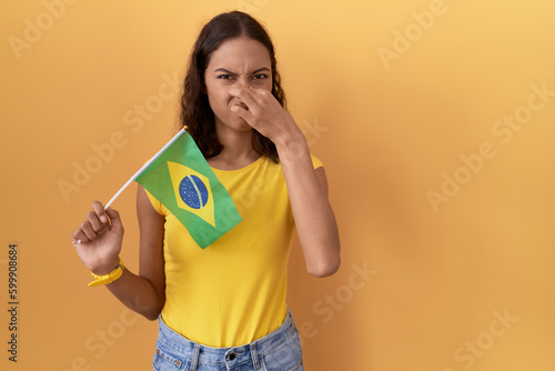Young hispanic woman holding brazil flag smelling something stinky and disgusting  intolerable smell  holding breath with fingers on nose. bad smell