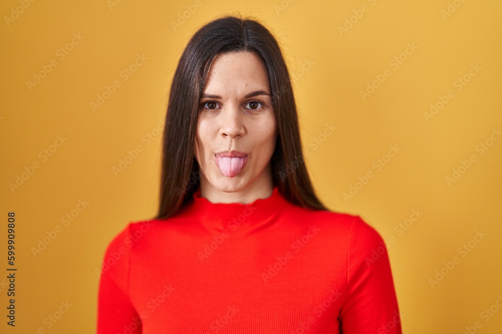 Fototapeta premium Young hispanic woman standing over yellow background sticking tongue out happy with funny expression. emotion concept.