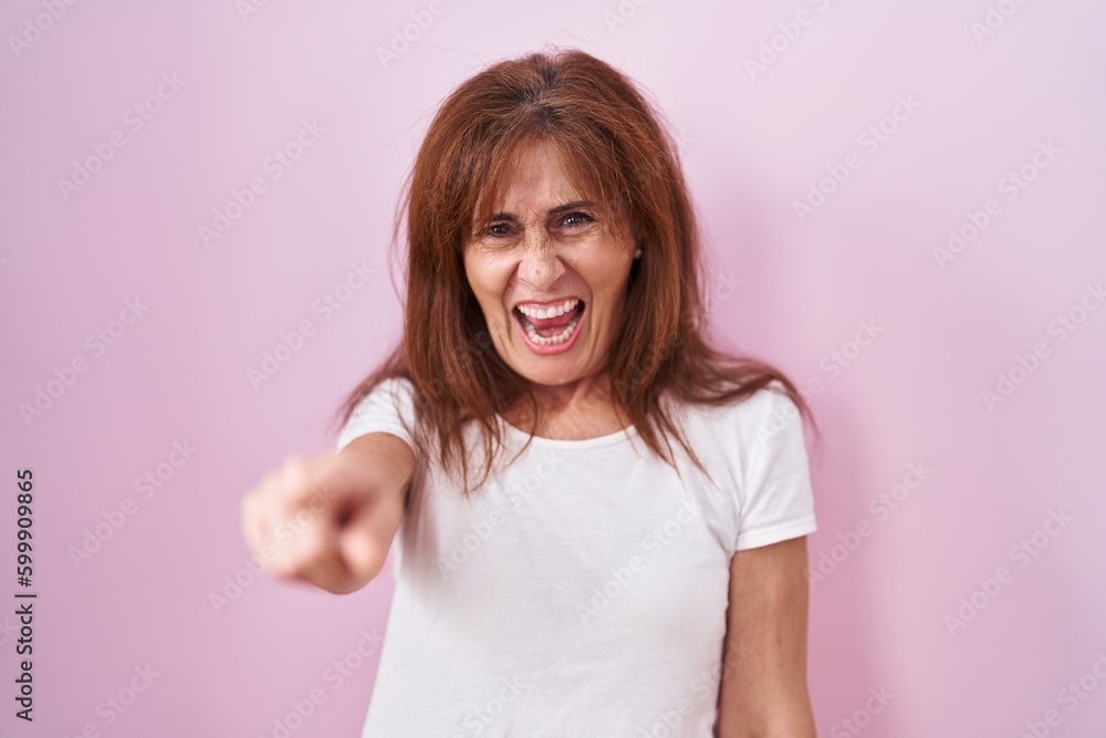Middle age woman standing over pink background pointing displeased and frustrated to the camera, angry and furious with you Middle age woman standing over pink background pointing displeased and frustrated to the camera, angry and furious with you