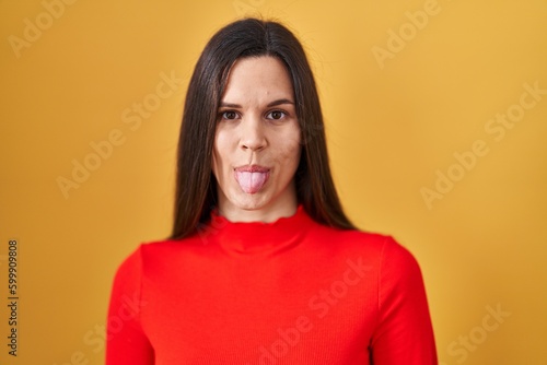 Young hispanic woman standing over yellow background sticking tongue out happy with funny expression. emotion concept.