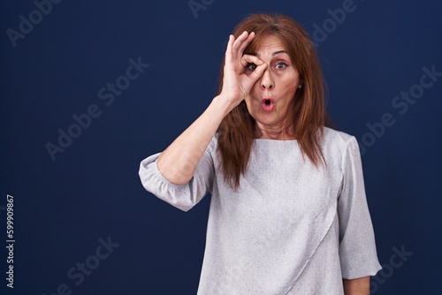 Middle age woman standing over blue background doing ok gesture shocked with surprised face, eye looking through fingers. unbelieving expression.