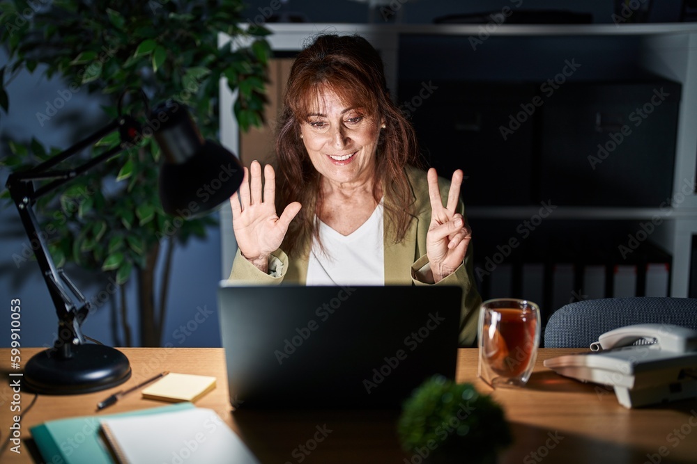 Middle age hispanic woman working using computer laptop at night showing and pointing up with fingers number seven while smiling confident and happy. Middle age hispanic woman working using computer laptop at night showing and pointing up with fingers number seven while smiling confident and happy.