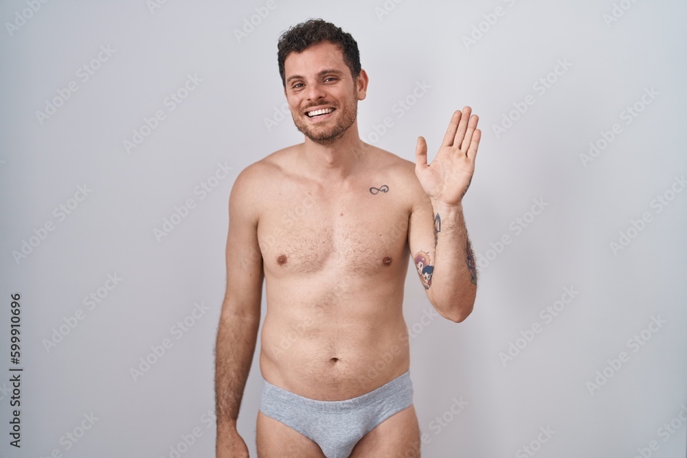 Young hispanic man standing shirtless wearing underware waiving saying hello happy and smiling, friendly welcome gesture Young hispanic man standing shirtless wearing underware waiving saying hello happy and smiling, friendly welcome gesture