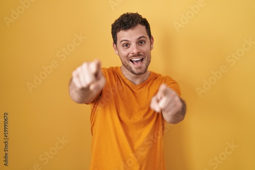 Young hispanic man standing over yellow background pointing to you and the camera with fingers, smiling positive and cheerful