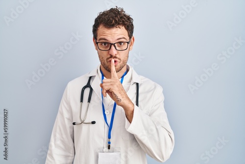 Young hispanic man wearing doctor uniform and stethoscope asking to be quiet with finger on lips. silence and secret concept.