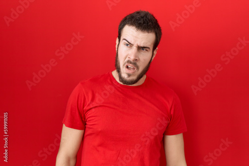 Young hispanic man wearing casual red t shirt in shock face  looking skeptical and sarcastic  surprised with open mouth