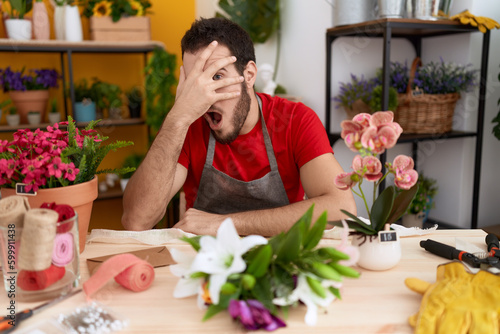 Young hispanic man working at florist shop peeking in shock covering face and eyes with hand  looking through fingers afraid