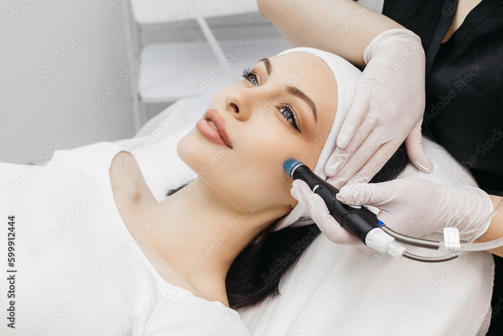 View of doctor cosmetologist doing anti aging procedure in cosmetology office. Satisfied woman in disposable hat lying on couch and relaxing. Working with Apparatus. View of doctor cosmetologist doing anti aging procedure in cosmetology office. Satisfied woman in disposable hat lying on couch and relaxing. Working with Apparatus.