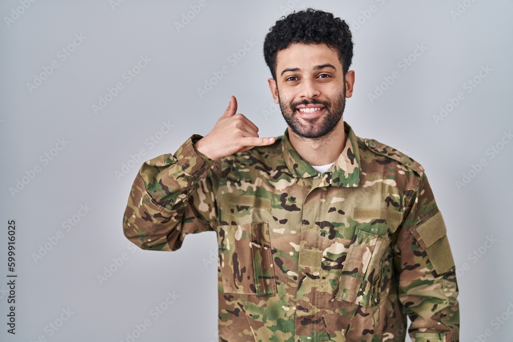 Arab man wearing camouflage army uniform smiling doing phone gesture with hand and fingers like talking on the telephone. communicating concepts.
