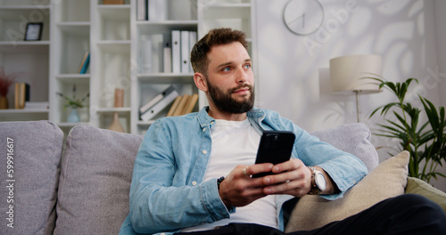 Portrait of attractive young man sitting on the sofa using phone smile in the modern apartment texting message content on smartphone social media applications online scrolling tapping technology.
