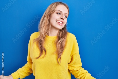 Young caucasian woman standing over blue background smiling cheerful with open arms as friendly welcome, positive and confident greetings