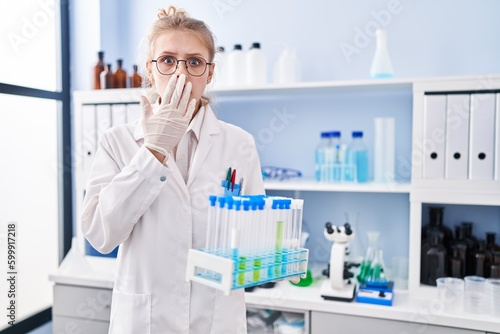 Young caucasian woman working at scientist laboratory holding samples covering mouth with hand, shocked and afraid for mistake. surprised expression