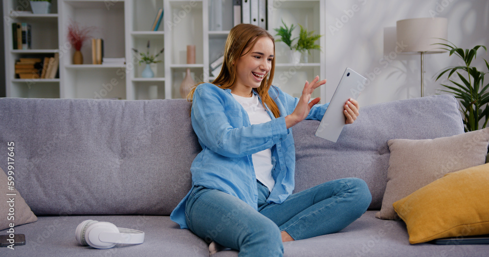 Obraz premium Pretty happy smiling Caucasian young woman sitting on comfortable sofa, using digital computer tablet internet study work shopping online using social media apps or playing game at home.