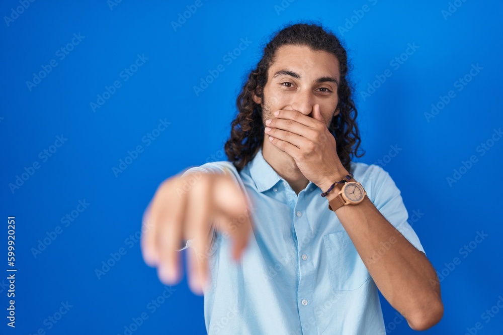 Young hispanic man standing over blue background laughing at you, pointing finger to the camera with hand over mouth, shame expression Young hispanic man standing over blue background laughing at you, pointing finger to the camera with hand over mouth, shame expression