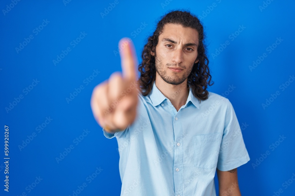 Young hispanic man standing over blue background pointing with finger up and angry expression, showing no gesture Young hispanic man standing over blue background pointing with finger up and angry expression, showing no gesture