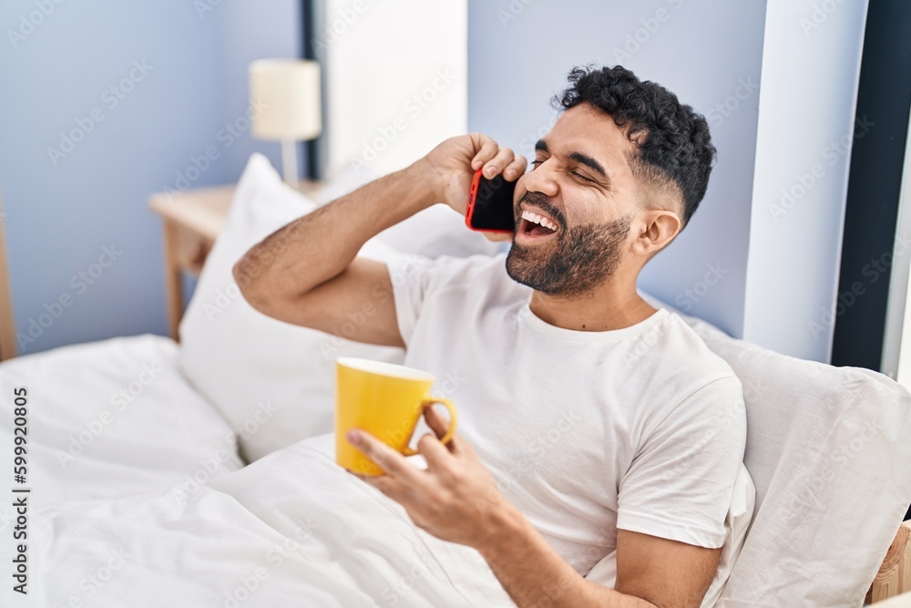 Young hispanic man talking on smartphone drinking coffee sitting on bed at bedroom Young hispanic man talking on smartphone drinking coffee sitting on bed at bedroom
