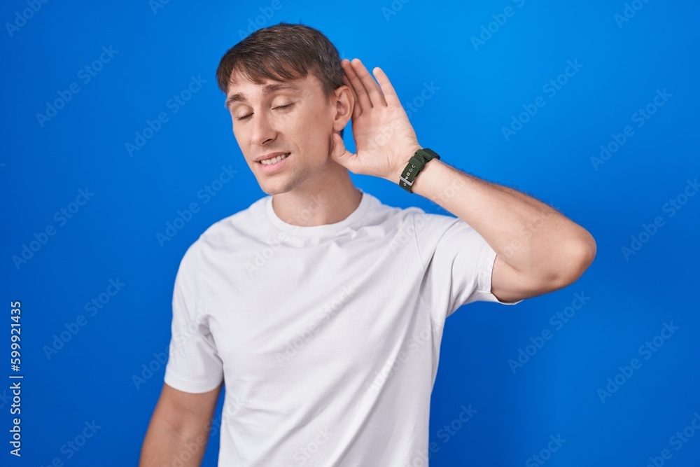 Caucasian blond man standing over blue background smiling with hand over ear listening an hearing to rumor or gossip. deafness concept. Caucasian blond man standing over blue background smiling with hand over ear listening an hearing to rumor or gossip. deafness concept.