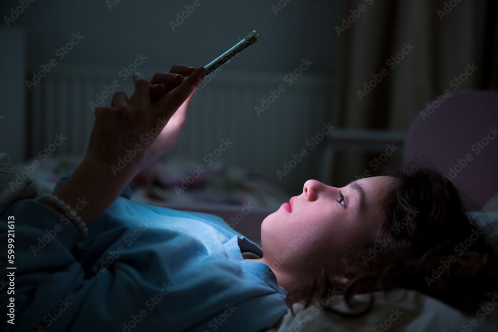 Obraz premium A child using smart phone lying in bed late at night, playing games, watching videos online, scrolling screen. Children's screen addiction and parent control concept. Child's room at night.