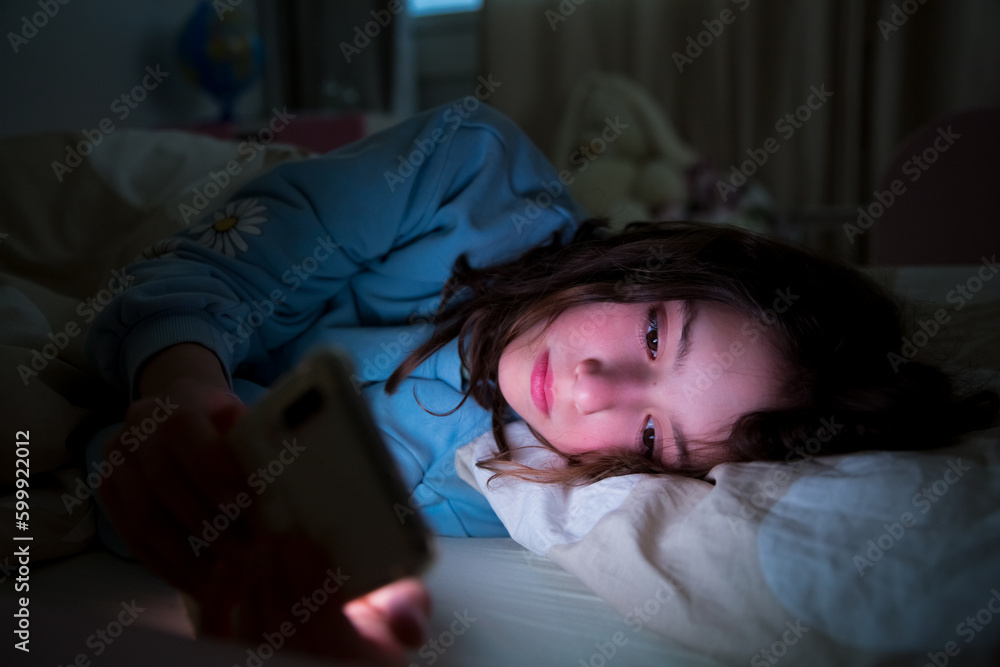 A child using smart phone lying in bed late at night, playing games, watching videos online, scrolling screen. Children's screen addiction and parent control concept. Child's room at night. A child using smart phone lying in bed late at night, playing games, watching videos online, scrolling screen. Children's screen addiction and parent control concept. Child's room at night.