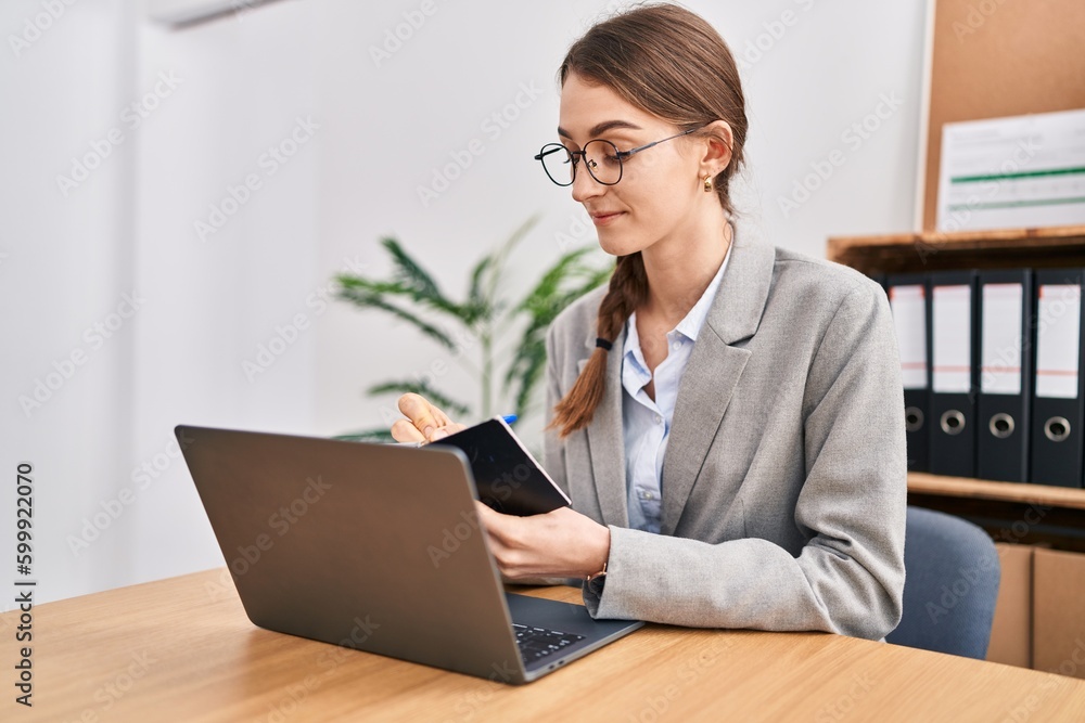 Young caucasian woman business worker using laptop writing on notebook at office Young caucasian woman business worker using laptop writing on notebook at office