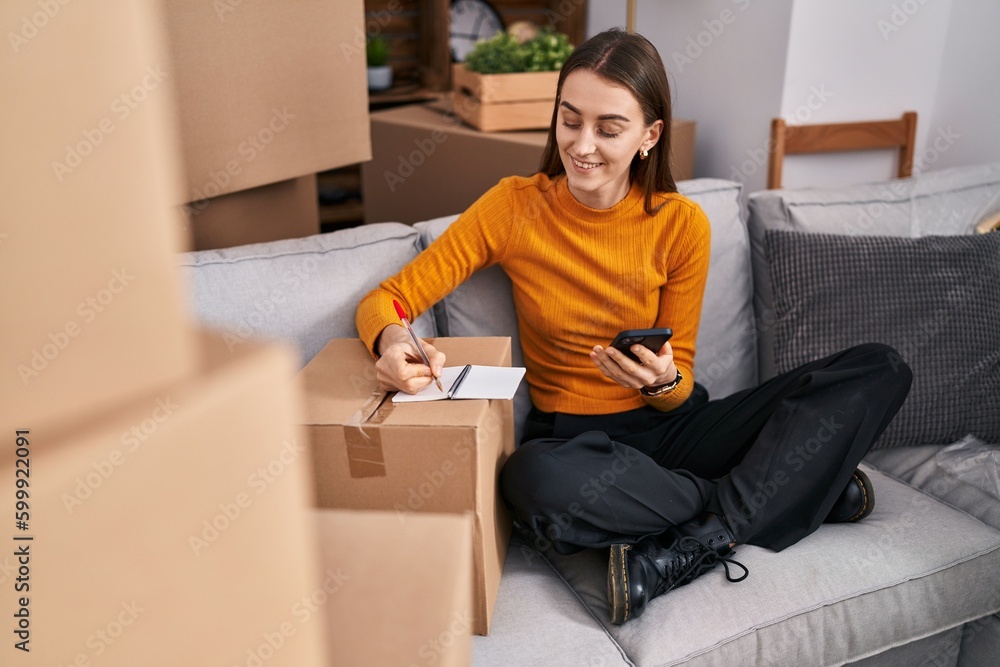 Young caucasian woman using smartphone writing on notebook at new home Young caucasian woman using smartphone writing on notebook at new home