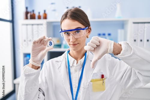 Young caucasian woman working at scientist laboratory holding bitcoin with angry face  negative sign showing dislike with thumbs down  rejection concept