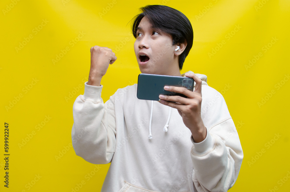 Happy expressive handsome young male gamer playing online game on smartphone isolated on yellow background