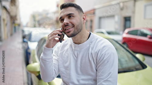 Young hispanic man talking on smartphone sitting on car at street