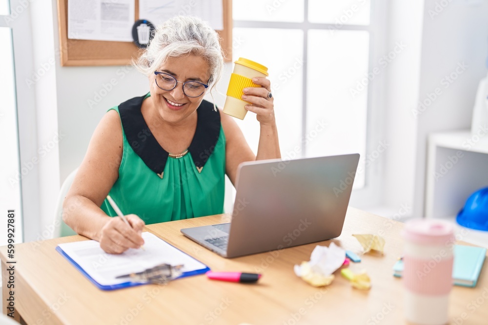 Middle age grey-haired woman business worker writing on document drinking coffee at office Middle age grey-haired woman business worker writing on document drinking coffee at office