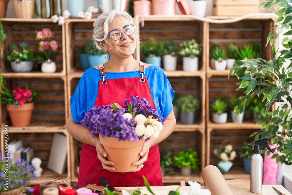 Middle age woman with grey hair working at florist shop holding plant winking looking at the camera with sexy expression, cheerful and happy face.