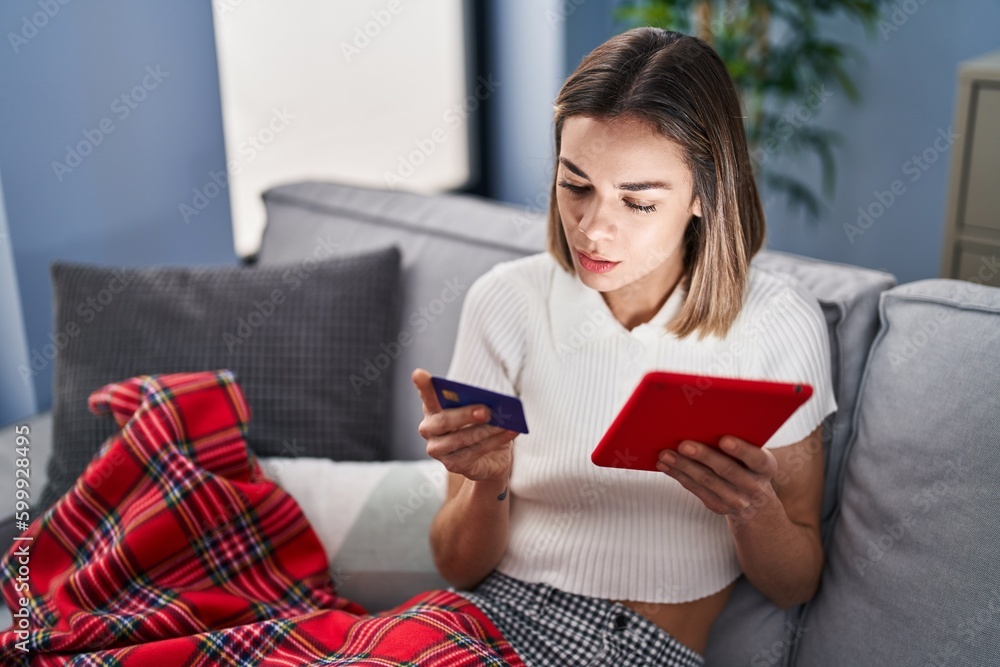 Young beautiful hispanic woman using touchpad and credit card sitting on sofa at home