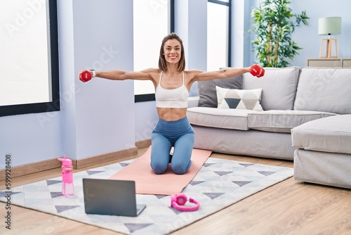 Young beautiful hispanic woman smiling confident having online dumbbells exercise at home